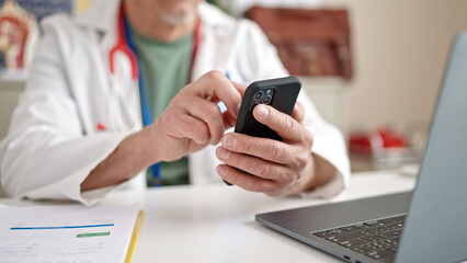Middle age grey-haired man doctor using smartphone and laptop working at clinic