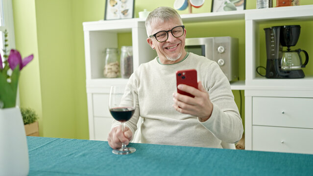 Middle age grey-haired man drinking glass of wine using smartphone at home - Powered by Adobe