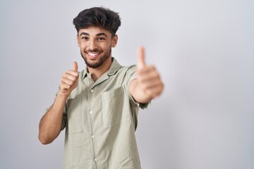 Fototapeta premium Arab man with beard standing over white background approving doing positive gesture with hand, thumbs up smiling and happy for success. winner gesture.