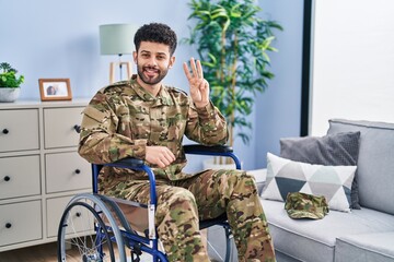 Fototapeta premium Arab man wearing camouflage army uniform sitting on wheelchair showing and pointing up with fingers number three while smiling confident and happy.
