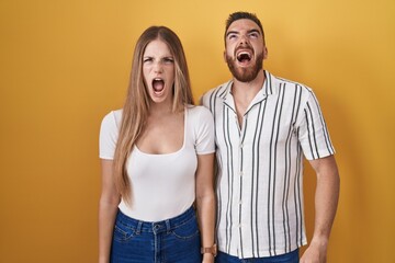 Young couple standing over yellow background angry and mad screaming frustrated and furious, shouting with anger. rage and aggressive concept.