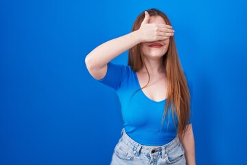 Redhead woman standing over blue background smiling and laughing with hand on face covering eyes for surprise. blind concept.