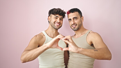 Two men couple hugging each other doing heart gesture over isolated pink background