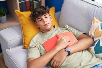 Young hispanic teenager holding book lying on sofa sleeping at home