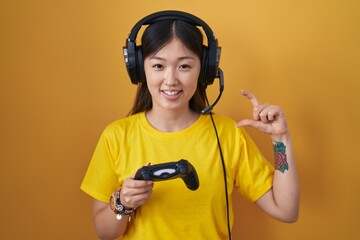 Chinese young woman playing video game holding controller smiling and confident gesturing with hand doing small size sign with fingers looking and the camera. measure concept.