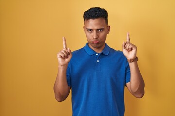 Young hispanic man standing over yellow background pointing up looking sad and upset, indicating...