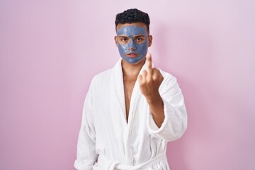 Young hispanic man wearing beauty face mask and bath robe showing middle finger, impolite and rude...