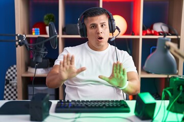 Young hispanic man playing video games moving away hands palms showing refusal and denial with afraid and disgusting expression. stop and forbidden.