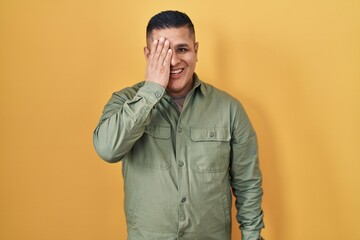 Hispanic young man standing over yellow background covering one eye with hand, confident smile on face and surprise emotion.