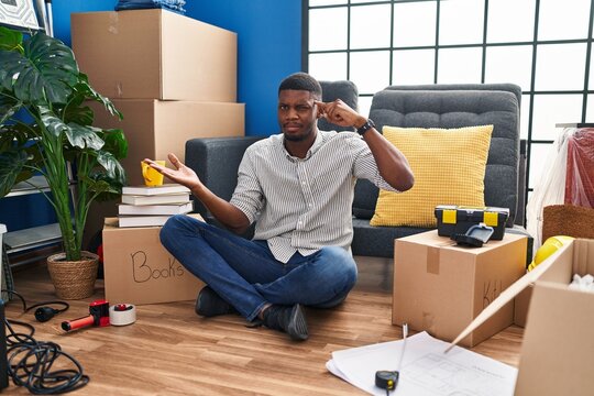 African american man sitting on the floor at new home confused and annoyed with open palm showing copy space and pointing finger to forehead. think about it.