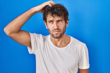 Hispanic young man standing over blue background confuse and wondering about question. uncertain with doubt, thinking with hand on head. pensive concept.