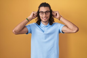 Young hispanic man standing over yellow background smiling pulling ears with fingers, funny gesture. audition problem