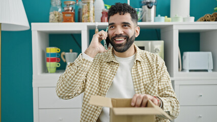 Young hispanic man talking on smartphone unpacking cardboard box at dinning room