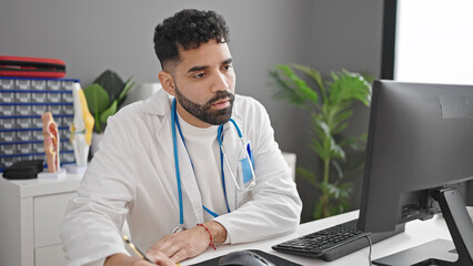 Young hispanic man doctor using computer writing notes at clinic