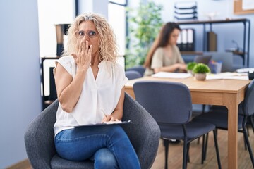 Middle age woman working at the office holding clipboard covering mouth with hand, shocked and...