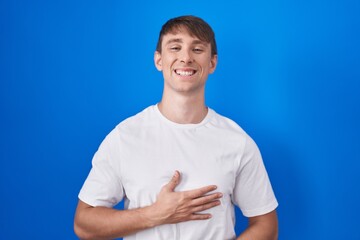 Caucasian blond man standing over blue background smiling and laughing hard out loud because funny crazy joke with hands on body.