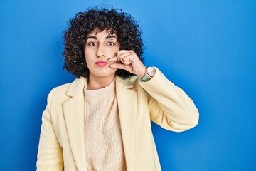 Young brunette woman with curly hair standing over blue background mouth and lips shut as zip with...
