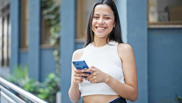 Young beautiful hispanic woman using smartphone smiling at street
