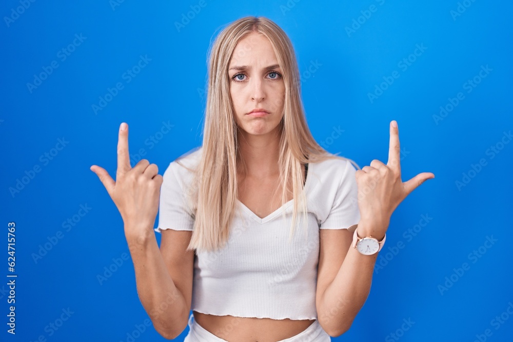 Poster young caucasian woman standing over blue background pointing up looking sad and upset, indicating di