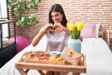 Obraz na płótnie Canvas Brunette young woman eating breakfast sitting on the bed smiling in love showing heart symbol and shape with hands. romantic concept.