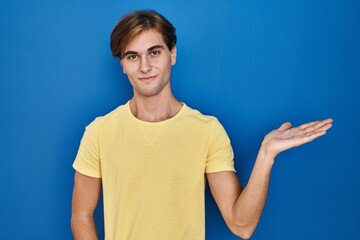 Young man standing over blue background smiling cheerful presenting and pointing with palm of hand looking at the camera.