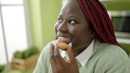 Fototapeta na wymiar African woman with braided hair eating cookie sitting on table at dinning room