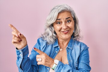 Obraz premium Middle age woman with grey hair standing over pink background smiling and looking at the camera pointing with two hands and fingers to the side.