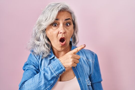 Middle age woman with grey hair standing over pink background surprised pointing with finger to the side, open mouth amazed expression.
