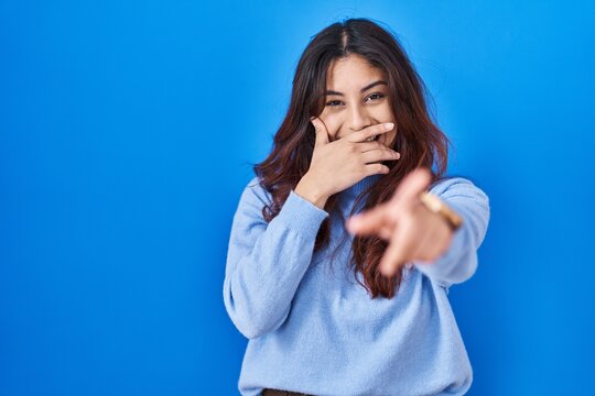 Hispanic young woman standing over blue background laughing at you, pointing finger to the camera with hand over mouth, shame expression