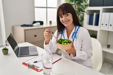 Young beautiful hispanic woman doctor using laptop eating salad at clinic
