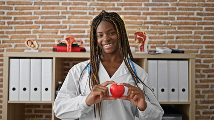 African american woman doctor smiling confident holding heart at the clinic