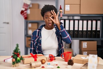 African american woman working at small business doing christmas decoration doing ok gesture shocked with surprised face, eye looking through fingers. unbelieving expression.