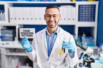 Young hispanic man working at scientist laboratory holding your donation matters holding blood sample winking looking at the camera with sexy expression, cheerful and happy face.