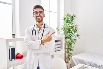 Hispanic man wearing doctor uniform and stethoscope smiling cheerful pointing with hand and finger up to the side