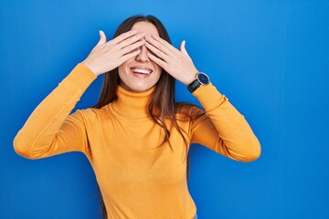 Young brunette woman standing over blue background covering eyes with hands smiling cheerful and funny. blind concept.