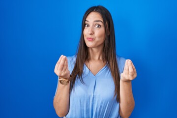 Fototapeta premium Young brunette woman standing over blue background doing money gesture with hands, asking for salary payment, millionaire business