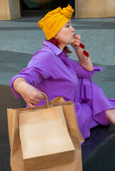woman with craft shopping bags in the city. The concept of fashionable Consumerism and ecology. An adult Asian woman in a purple suit with red and yellow accents enjoys shopping . a sunny day.