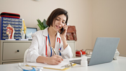 Young caucasian woman doctor talking on phone writing on document at clinic