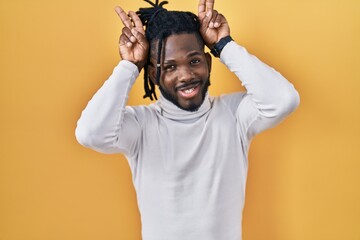 African man with dreadlocks wearing turtleneck sweater over yellow background posing funny and crazy with fingers on head as bunny ears, smiling cheerful