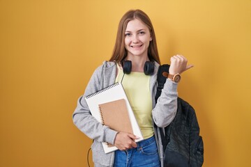 Young caucasian woman wearing student backpack and holding books pointing to the back behind with hand and thumbs up, smiling confident
