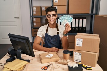 Young hispanic man working at small business ecommerce holding brazilian reals looking positive and happy standing and smiling with a confident smile showing teeth