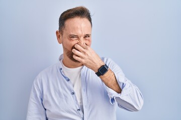 Middle age caucasian man standing over blue background smelling something stinky and disgusting, intolerable smell, holding breath with fingers on nose. bad smell