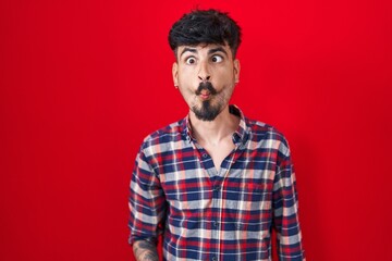 Young hispanic man with beard standing over red background making fish face with lips, crazy and comical gesture. funny expression.