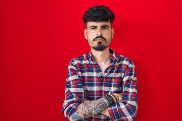 Young hispanic man with beard standing over red background skeptic and nervous, disapproving expression on face with crossed arms. negative person.