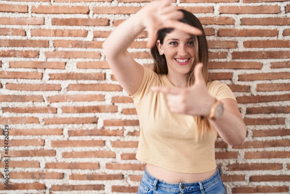 Sticker Young brunette woman standing over bricks wall smiling making frame with hands and fingers with happy face. creativity and photography concept.