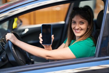 Obraz na płótnie Canvas Young hispanic woman showing screen smartphone sitting on car at street