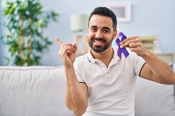 Young hispanic man with beard holding purple ribbon awareness smiling happy pointing with hand and...
