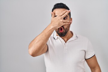 Young hispanic man with beard wearing casual clothes over white background peeking in shock covering face and eyes with hand, looking through fingers with embarrassed expression.