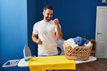 Young hispanic man with beard ironing clothes at home celebrating surprised and amazed for success with arms raised and eyes closed