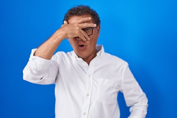 Middle age hispanic man standing over blue background peeking in shock covering face and eyes with hand, looking through fingers with embarrassed expression.